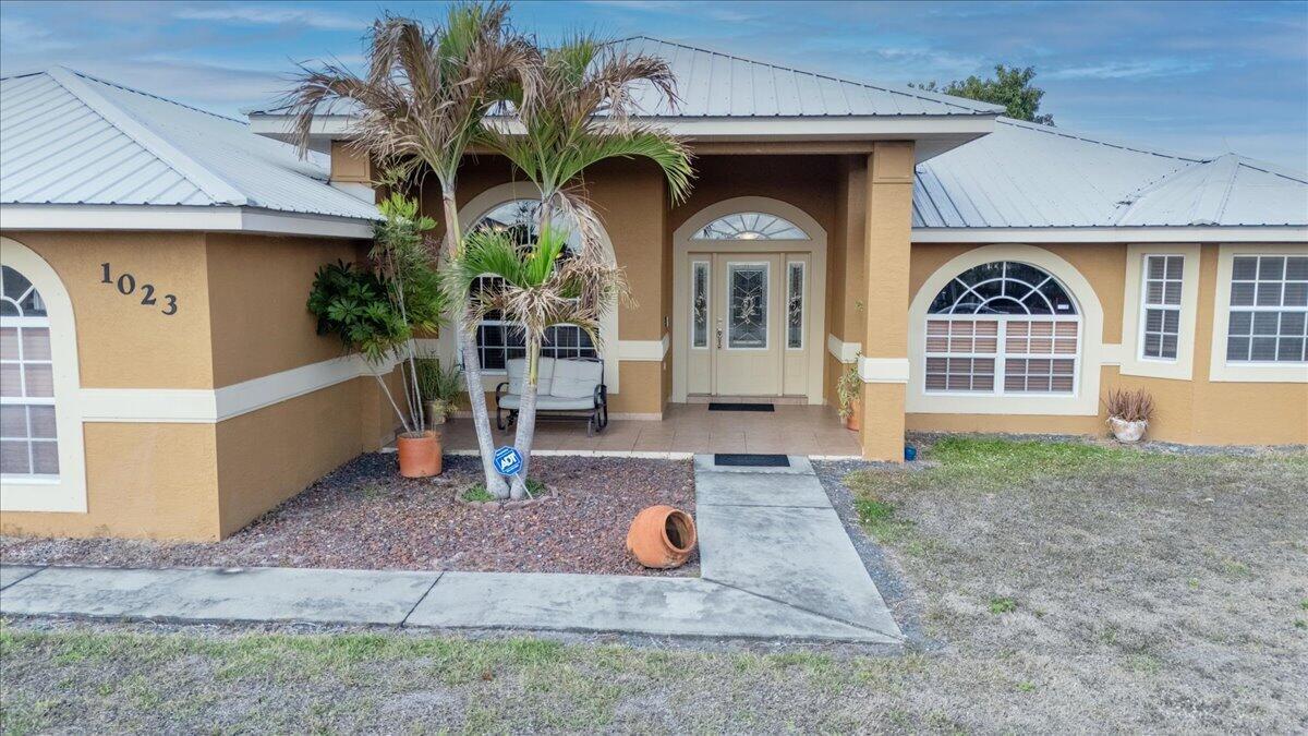 The front exterior of a tan house features a metal roof, arched windows, and palm trees — 1023 Caribbean Avenue, Clewiston FL 33440, CLEWISTON, FL
