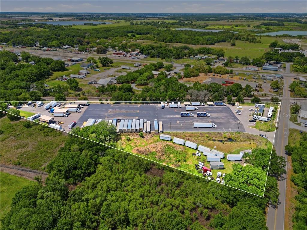 An aerial view shows a paved commercial lot with parked semi-trailers and surrounding wooded land — 100 S AIRPORT ROAD, LAKE WALES, FL