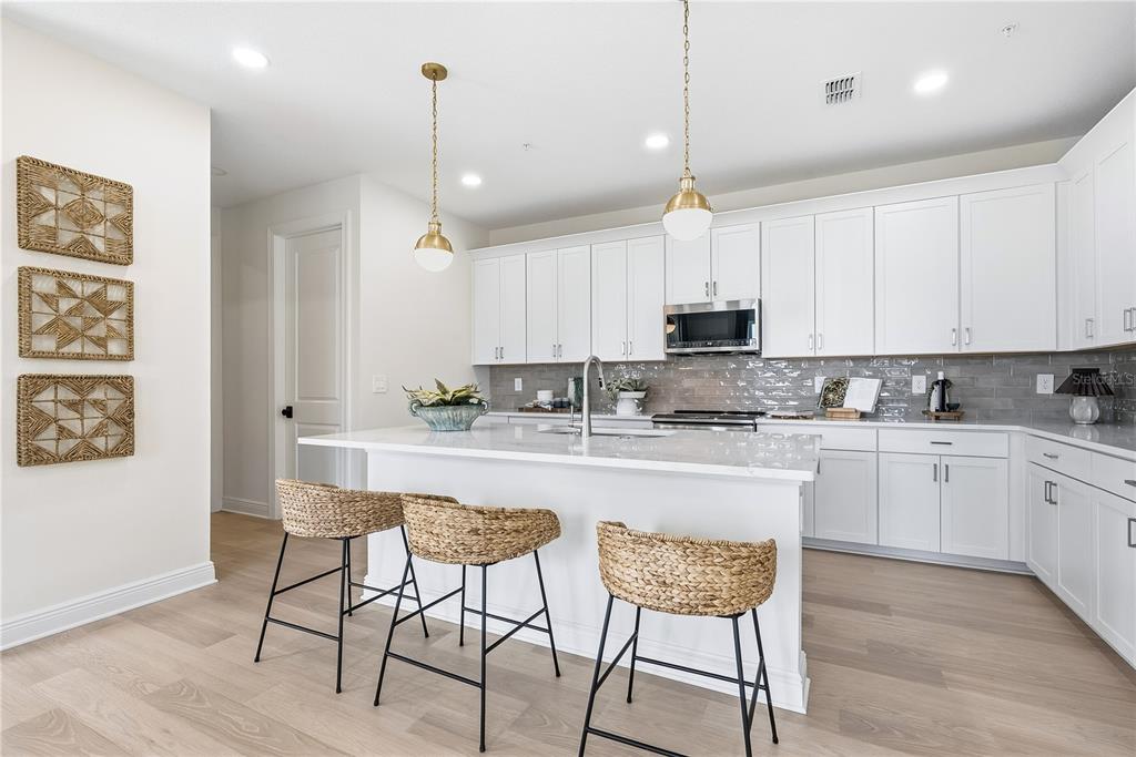 A modern kitchen features white cabinetry, a large island with three woven stools, and wood flooring — 17818 BROADLEAF LOOP, VENICE, FL