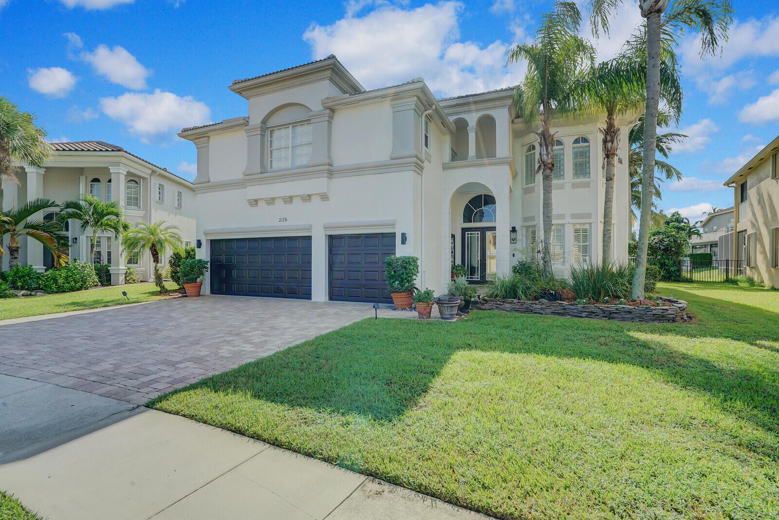 A two-story stucco house with a paved driveway, dark garage doors, and palm trees — 2176 Bellcrest Circle, Royal Palm Beach FL 33411, ROYAL PALM BEACH, FL
