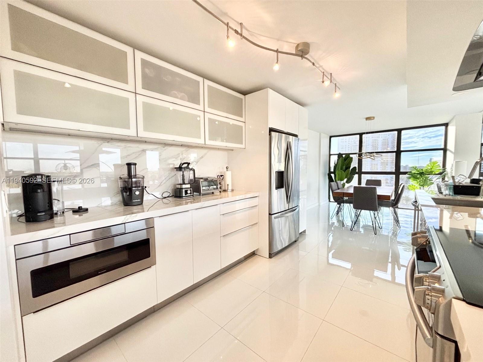 A modern kitchen features white cabinetry, stainless steel appliances, and a dining area with windows — 200 Leslie Dr # 831, Hallandale Beach FL 33009, HALLANDALE BEACH, FL