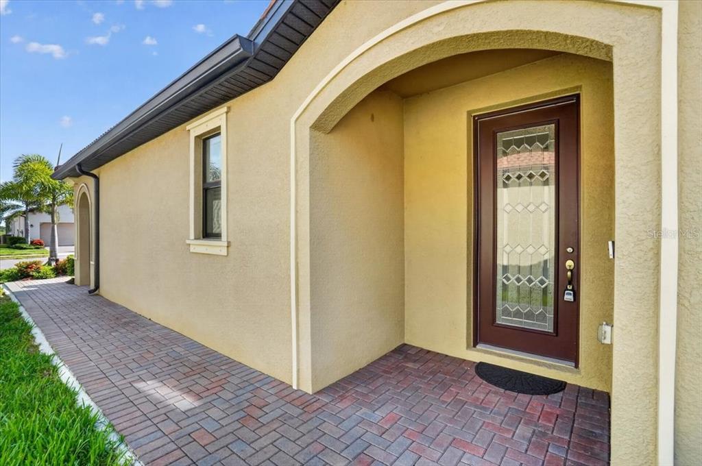 A tan stucco house exterior features a brick walkway leading to a brown glass-paneled front door — 24229 SPARTINA DRIVE, VENICE, FL