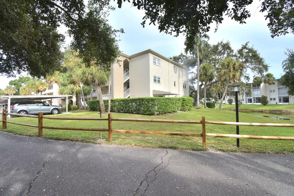A three-story apartment building sits behind a wooden fence next to a parking area — 940 VIRGINIA STREET UNIT 108, DUNEDIN, FL