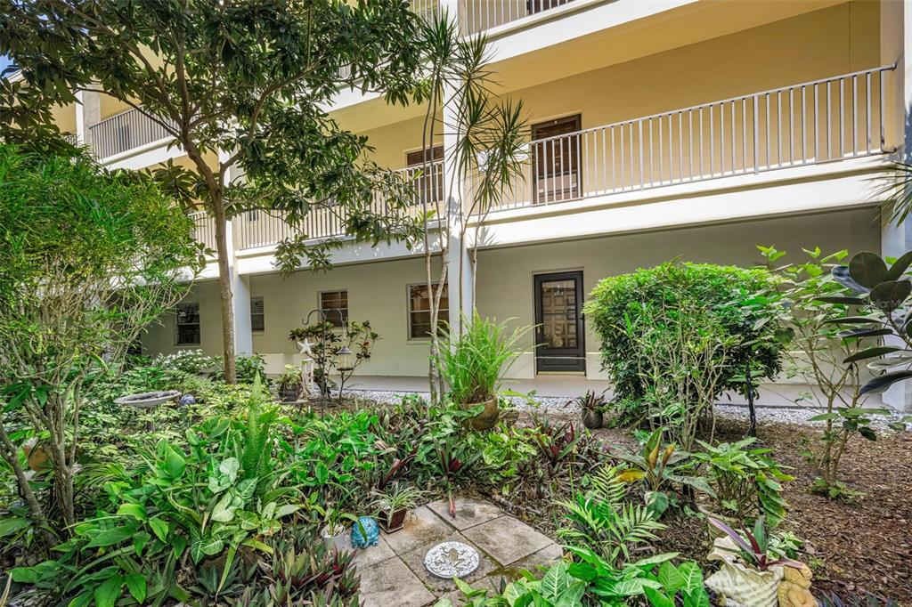A ground-level patio area features a dark door, white railings, and lush tropical landscaping — 2700 BAYSHORE BOULEVARD UNIT 6107, DUNEDIN, FL