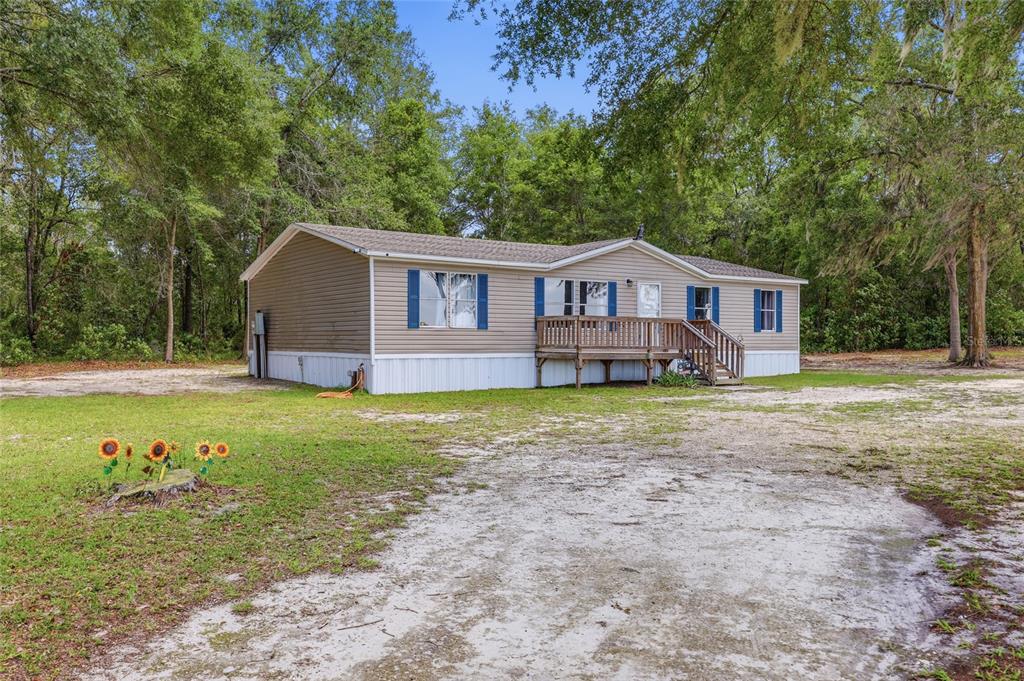 A tan manufactured home with blue shutters sits on a sandy lot surrounded by trees — 3562 NW 20TH AVENUE, BELL, FL