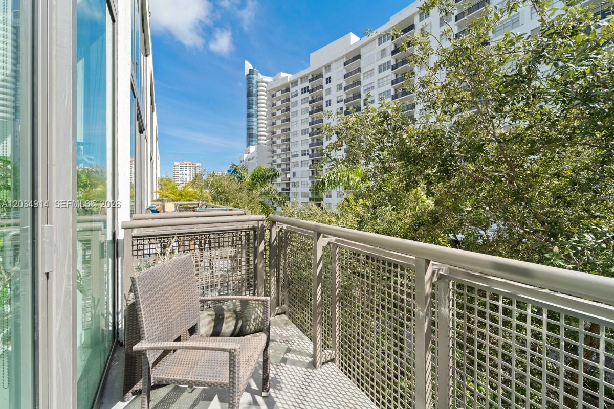 A balcony with a wicker chair and metal railing overlooks trees and nearby high-rise buildings — 6000 Collins Ave # 517, Miami Beach FL 33140, MIAMI BEACH, FL