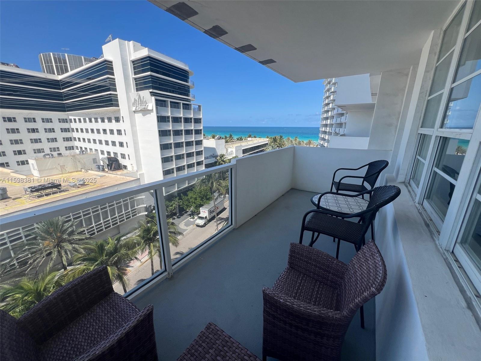 A balcony with wicker chairs and a small table overlooks a street and the ocean — 100 Lincoln Rd # 808, Miami Beach FL 33139, MIAMI BEACH, FL