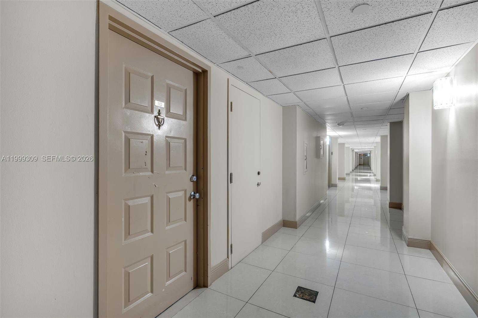 A long, brightly lit hallway features white tiled floors, a drop ceiling, and a beige door — 1201 S Ocean Dr # 110S, Hollywood FL 33019, HOLLYWOOD, FL