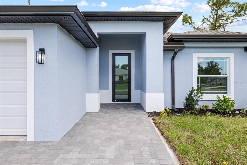 A light blue house exterior features a paved walkway leading to a modern glass door — 152 ALBATROSS ROAD, ROTONDA WEST, FL