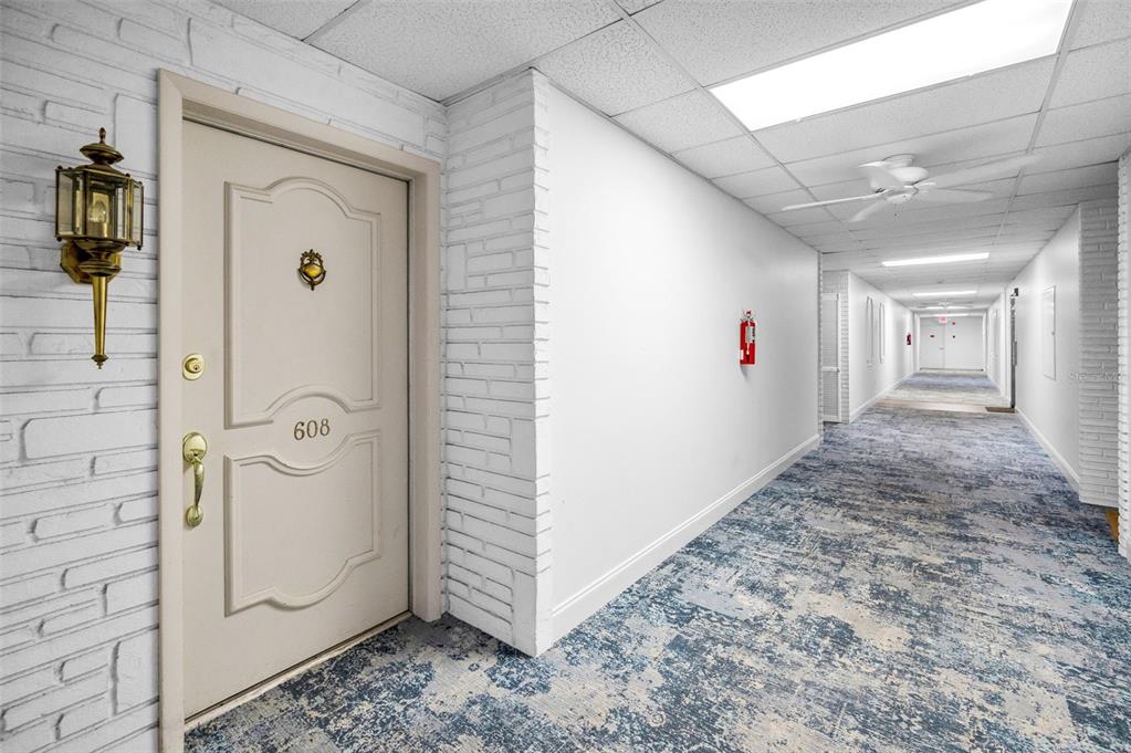 A hallway with a white door numbered 608, white brick walls, and patterned blue carpeting — 100 BLUFF VIEW DRIVE UNIT 608C, BELLEAIR BLUFFS, FL