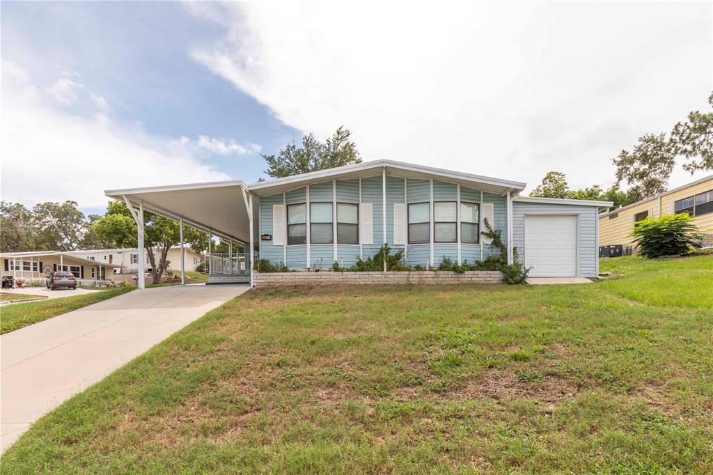 A light blue manufactured home features a front carport, a small garage, and a lawn — 7379 DEL RIO AVENUE, BROOKSVILLE, FL