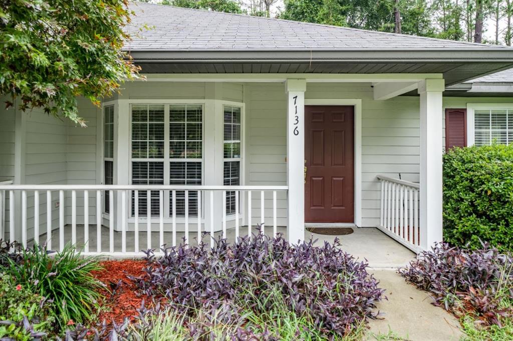 A light green house exterior features a covered porch, a red door, and purple landscaping — 7136 SW 81ST DRIVE, GAINESVILLE, FL