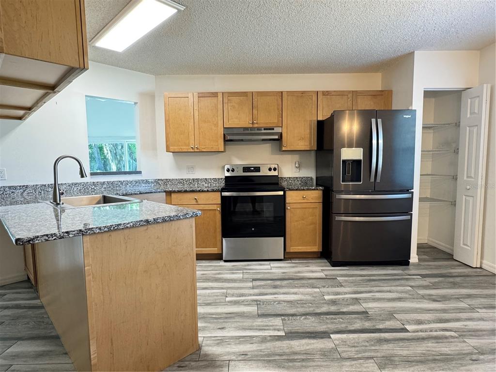 A kitchen features light wood cabinets, granite countertops, stainless steel appliances, and gray wood-look flooring — 215 FERNWOOD ST, LEESBURG, FL