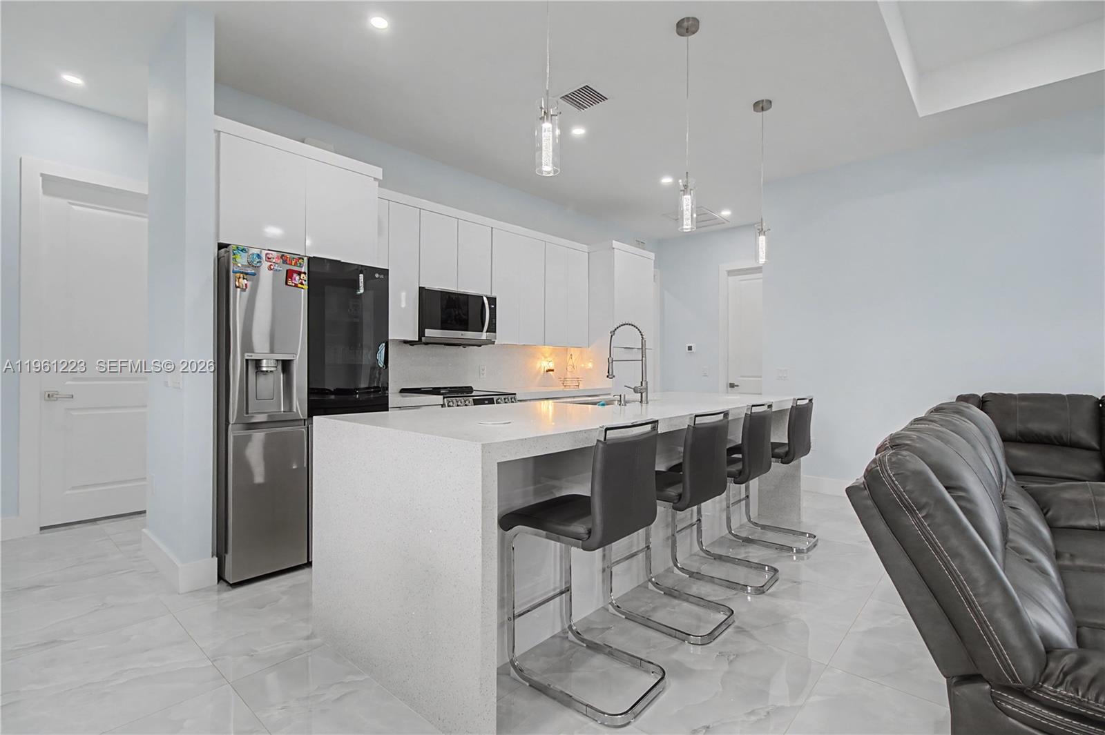 A modern kitchen features white cabinetry, a quartz waterfall island, and four gray bar stools — 3705 SW 23rd St, Lehigh Acres FL 33976, LEHIGH ACRES, FL