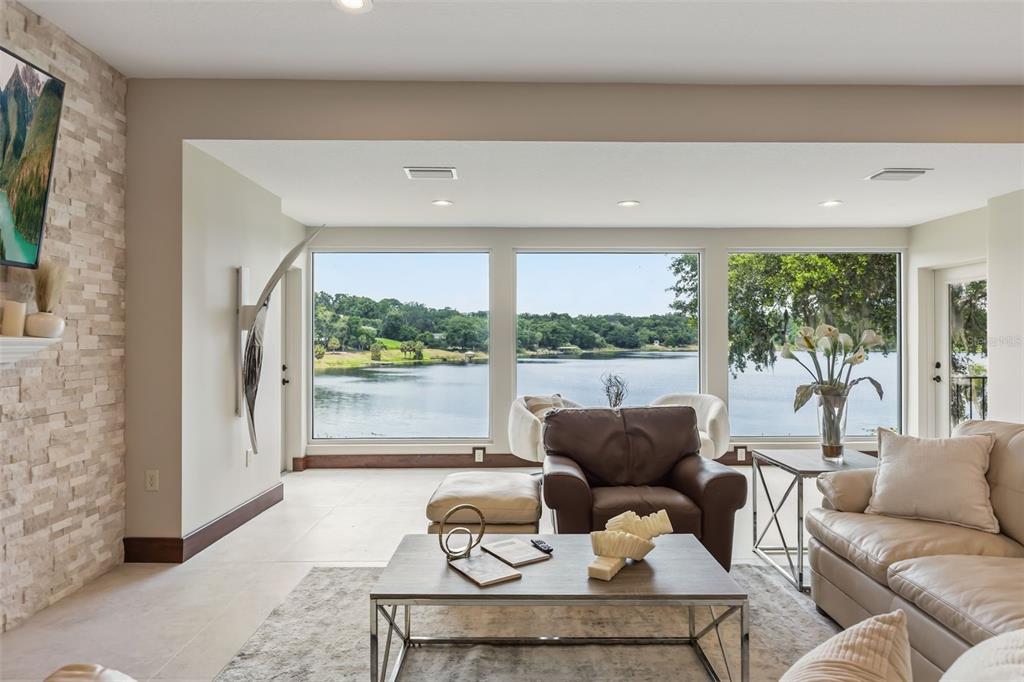 A living room features a stone fireplace, large windows overlooking a lake, and neutral furniture — 1909 COUNTRY CLUB DRIVE, EUSTIS, FL
