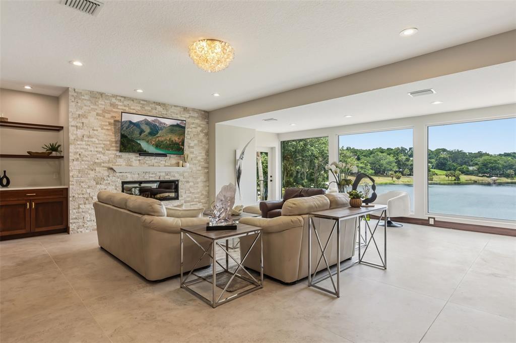 A living room features a stone fireplace, beige sofas, and large windows overlooking a lake — 1909 COUNTRY CLUB DRIVE, EUSTIS, FL