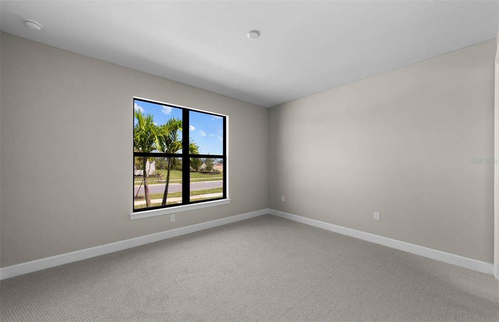 An empty bedroom features beige walls, light-colored carpeting, and a window overlooking a suburban street — 1626 PEPPER GRASS DRIVE SW, NORTH PORT, FL