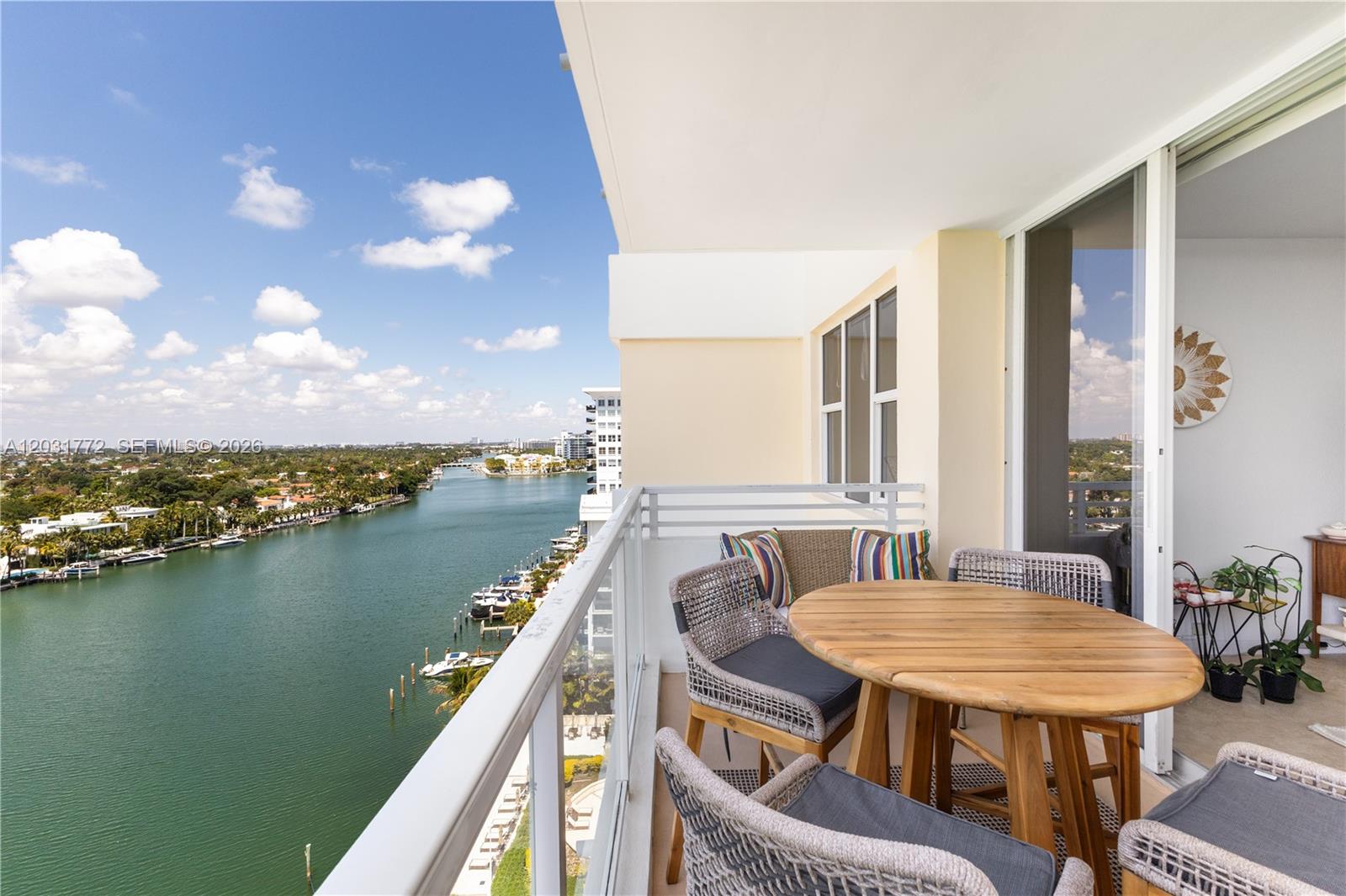 A balcony with a wooden table and chairs overlooks a river and coastal buildings — 5600 Collins Ave # 14R, Miami Beach FL 33140, MIAMI BEACH, FL