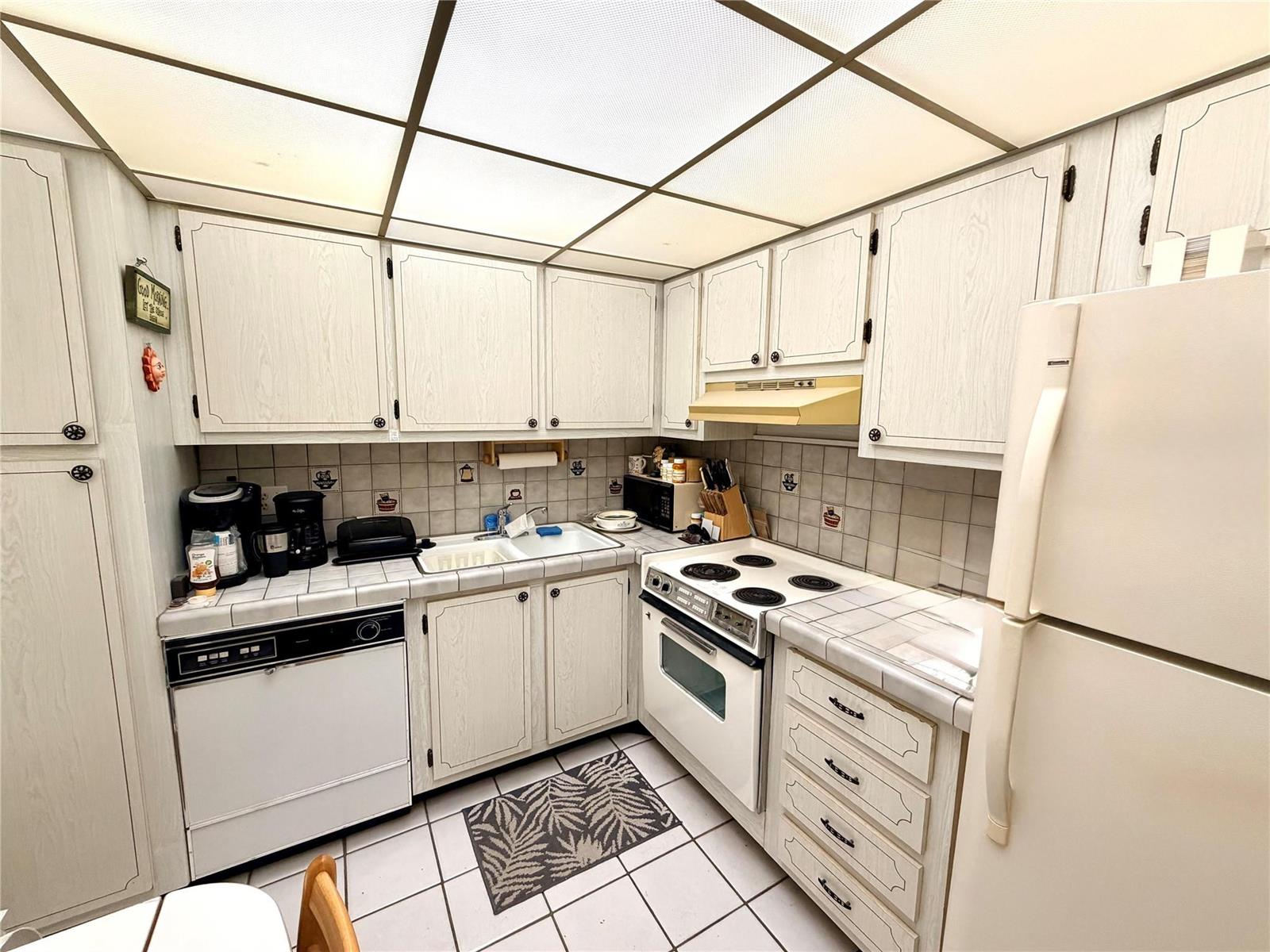 A kitchen features white wood-grain cabinets, tiled countertops, a drop ceiling, and white appliances — 136 S Cypress Road # 317, Pompano Beach FL 33060, POMPANO BEACH, FL