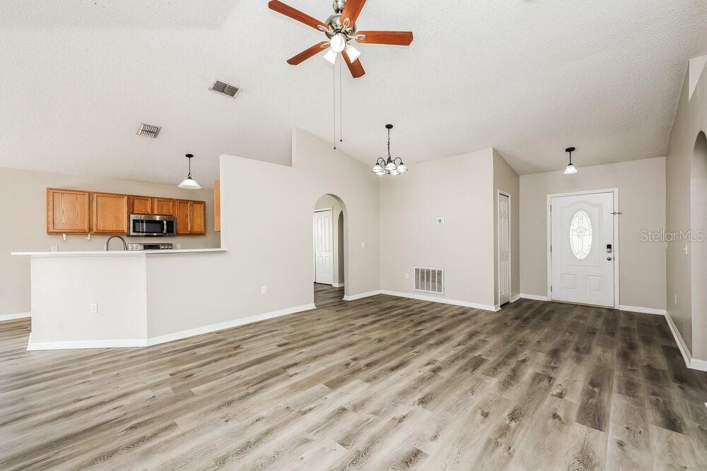 An open-concept living area features light wood-look flooring, a kitchen with wooden cabinets, and vaulted ceilings — 12938 BRIDLEFORD DRIVE, GIBSONTON, FL