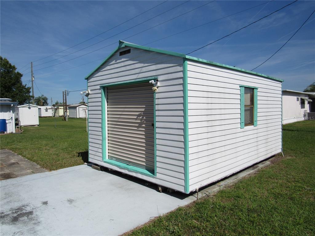 A white storage shed with a roll-up door and a single window sits on grass — 37241 CORA AVENUE, ZEPHYRHILLS, FL