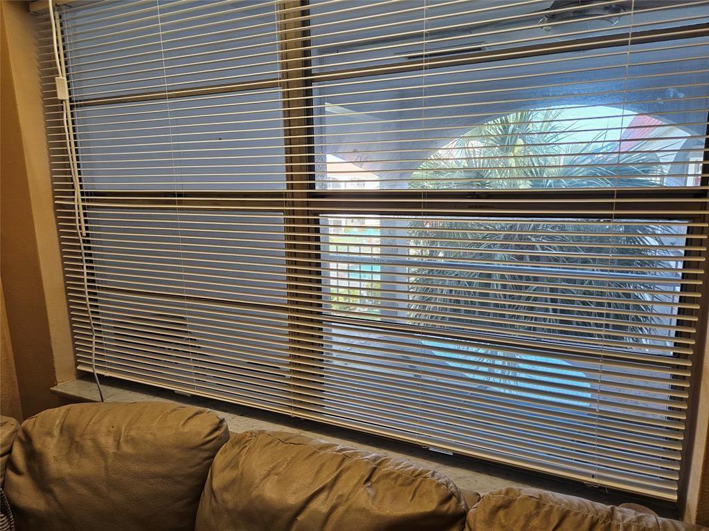 A window with horizontal blinds overlooks a patio area with a swimming pool and palm trees — 580 Egret Drive # 310, Hallandale Beach FL 33009, HALLANDALE BEACH, FL