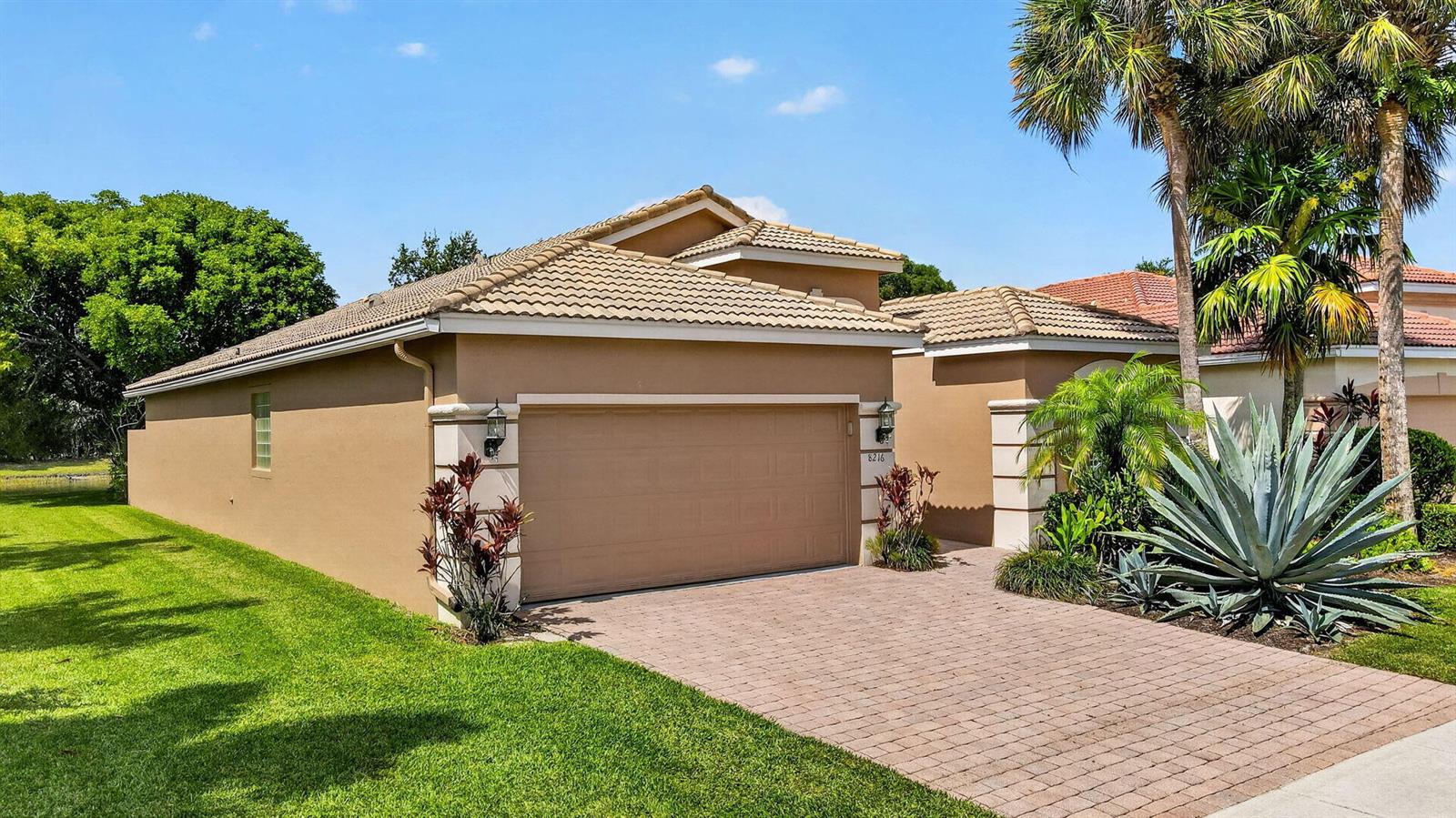 A tan single-story house features a two-car garage, a paver driveway, and tropical landscaping — 8216 Parini Way, Lake Worth FL 33467, LAKE WORTH, FL