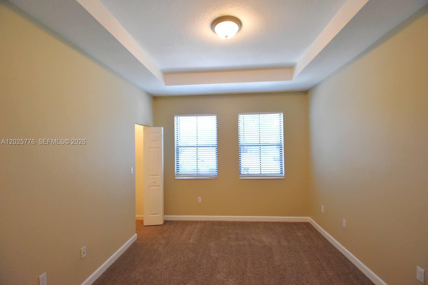 An empty bedroom features beige walls, carpeted flooring, two windows, and a tray ceiling — FORT LAUDERDALE, FL