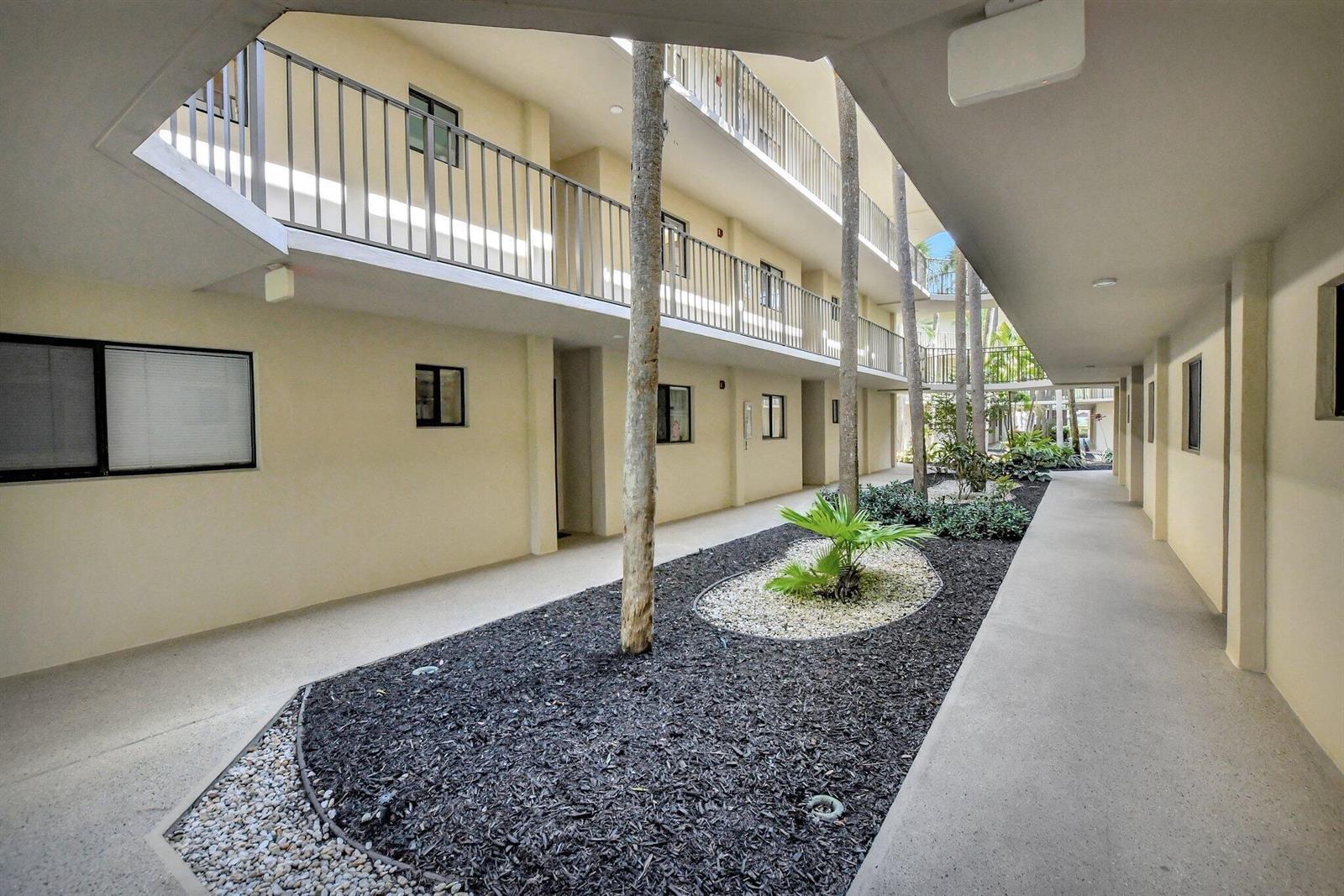 An open-air apartment complex courtyard features a central garden bed with trees and palm plants — 5340 Las Verdes Circle # 208, Delray Beach FL 33484, DELRAY BEACH, FL