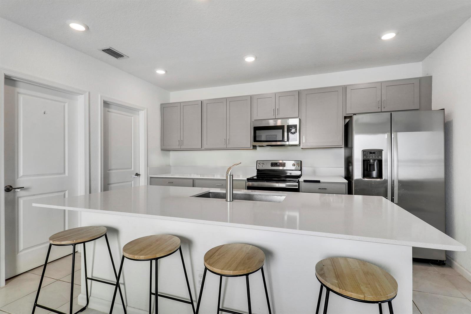 A modern kitchen features a white quartz island with bar stools, gray cabinetry, and stainless-steel appliances — 4233 Troon Place, Fort Pierce FL 34947, FORT PIERCE, FL