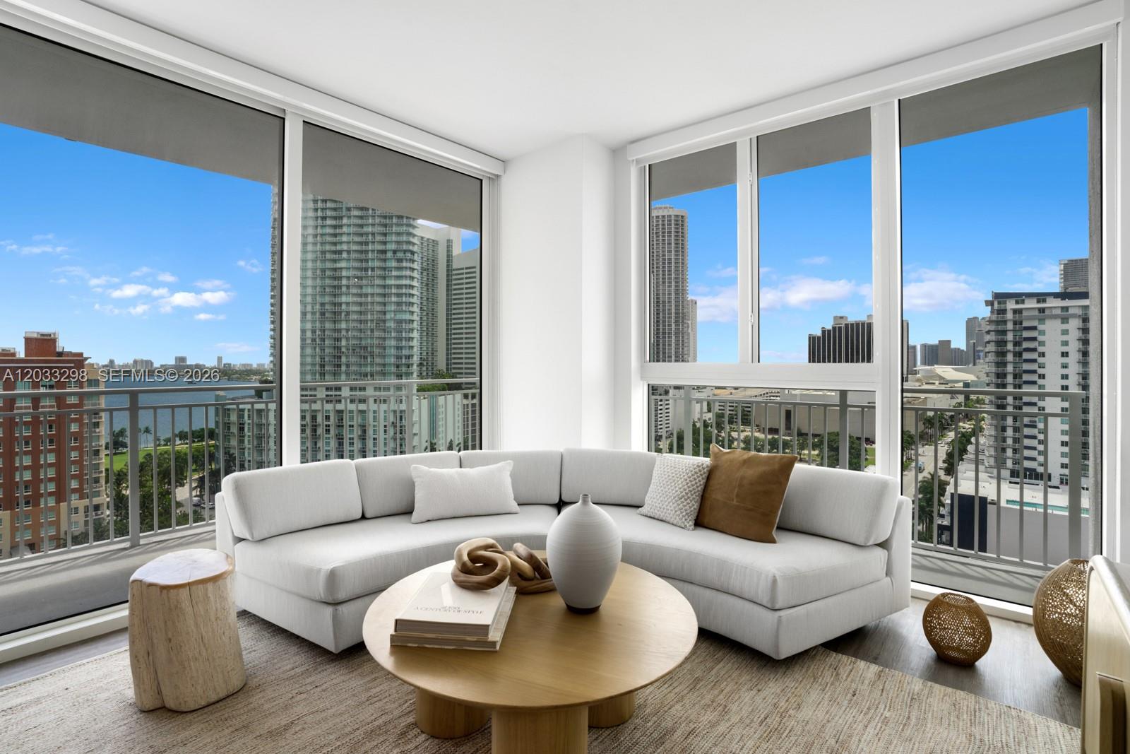 A living room features a curved white sofa, a round wooden coffee table, and floor-to-ceiling windows — 2000 Biscayne Blvd # 1009, Miami FL 33137, MIAMI, FL