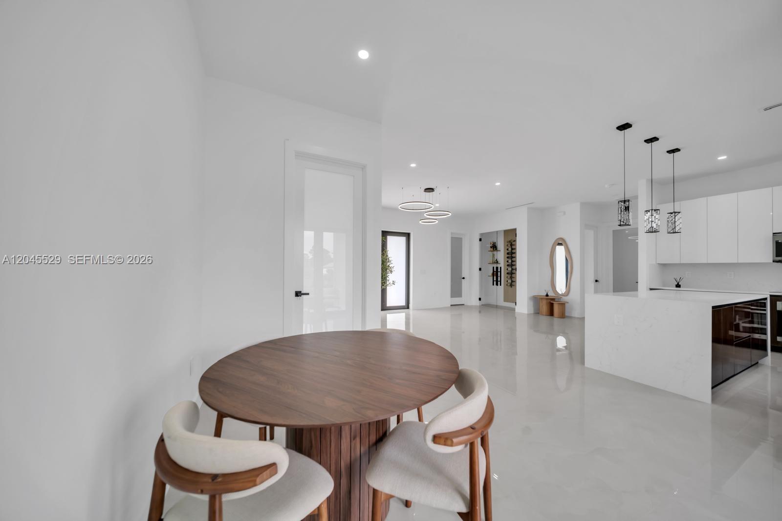 A modern dining area with a round wooden table opens into a white kitchen space — 323 LLOYD AVE S, Lehigh Acres FL 33974, LEHIGH ACRES, FL
