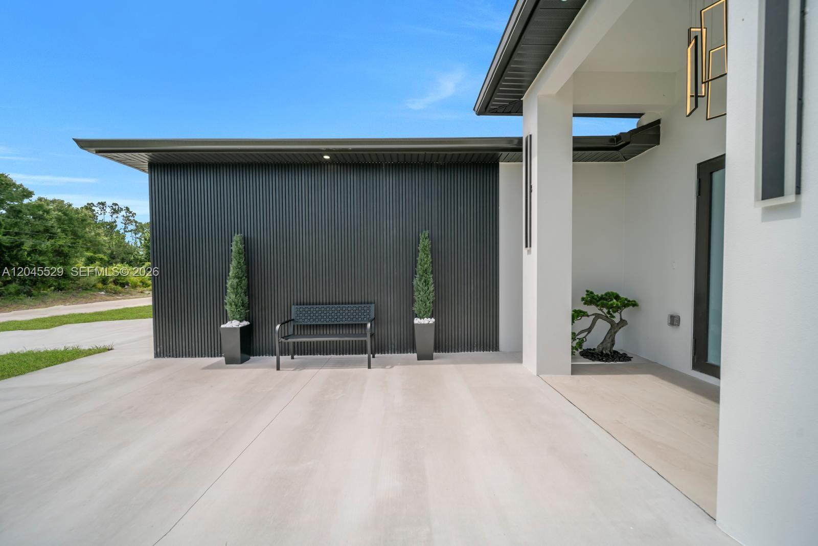 A modern home exterior features a black slatted wall, a metal bench, and potted topiaries — 323 LLOYD AVE S, Lehigh Acres FL 33974, LEHIGH ACRES, FL