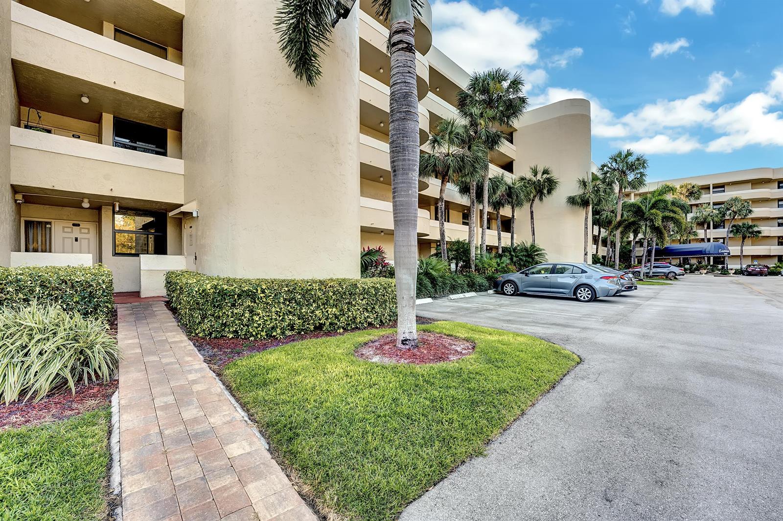 A multi-story beige apartment building with a paved walkway, palm trees, and parked cars — 1850 Homewood Boulevard # 1030, Delray Beach FL 33445, DELRAY BEACH, FL
