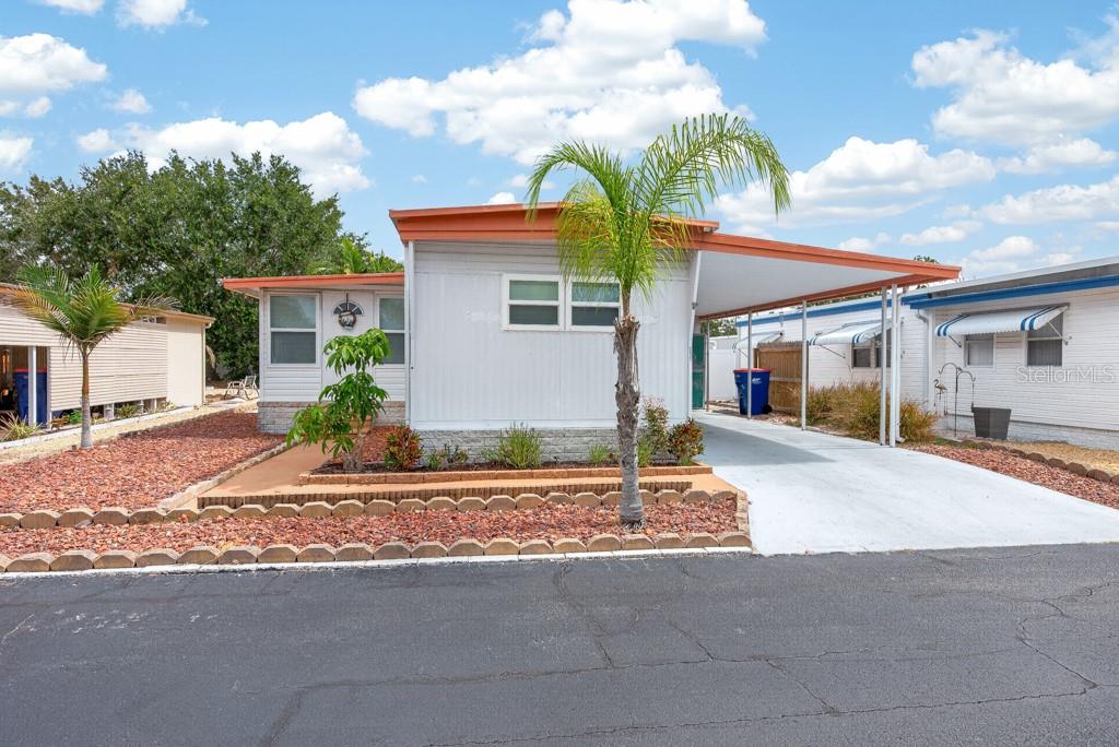 A single-story manufactured home with a carport, palm tree, and red rock landscaping — 3527 TIKI DRIVE, HOLIDAY, FL