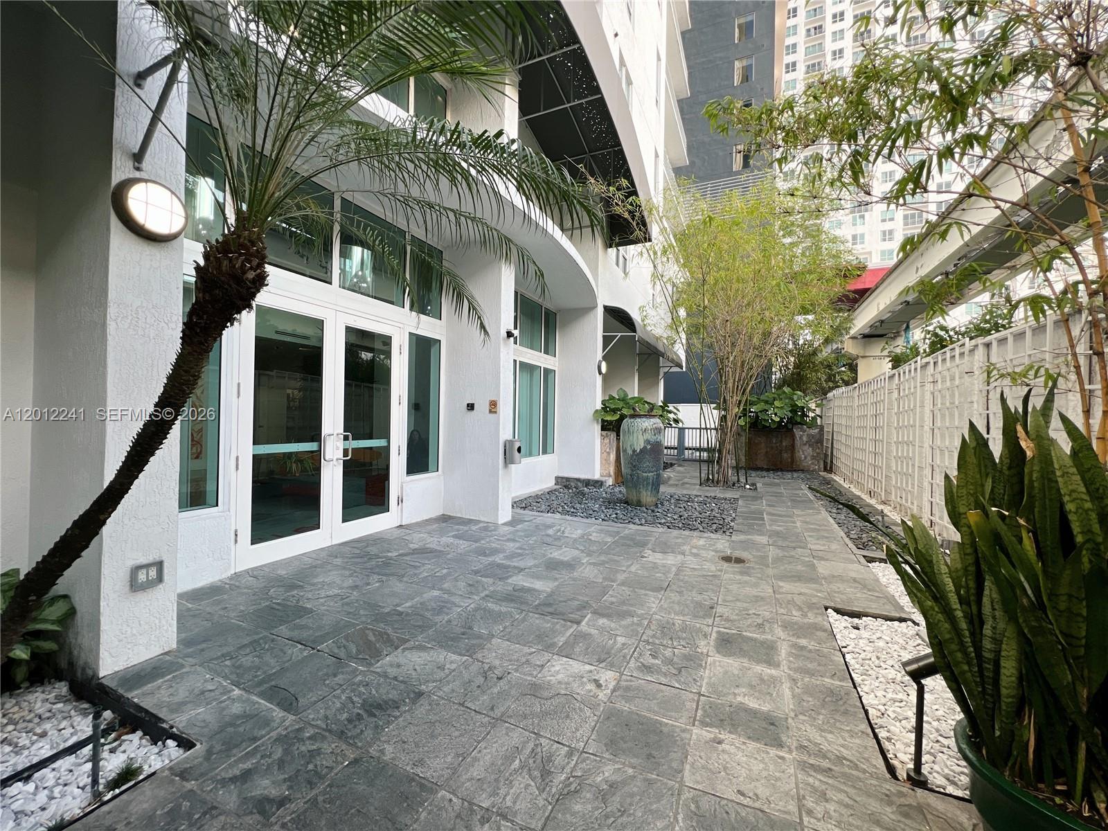 A paved patio area features a glass-paned entrance, potted plants, and a modern building exterior — 234 NE 3rd St # 703, Miami FL 33132, MIAMI, FL