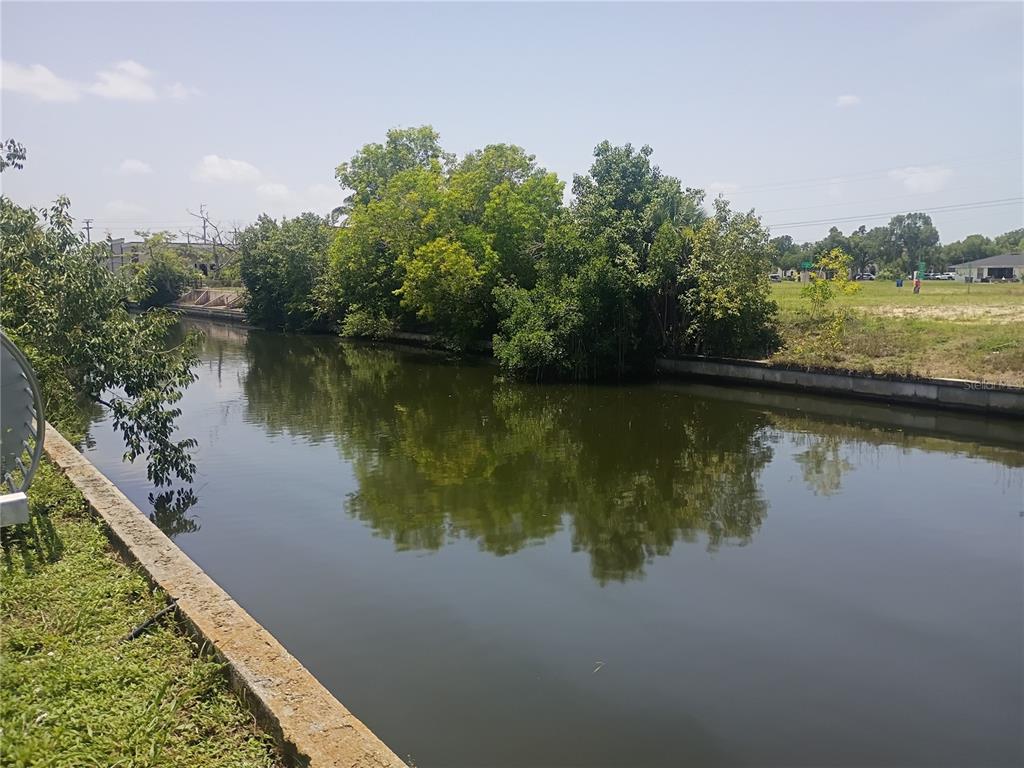 A concrete-lined canal flows past a cluster of trees and an open grassy lot — 918 SE 9TH LANE, CAPE CORAL, FL
