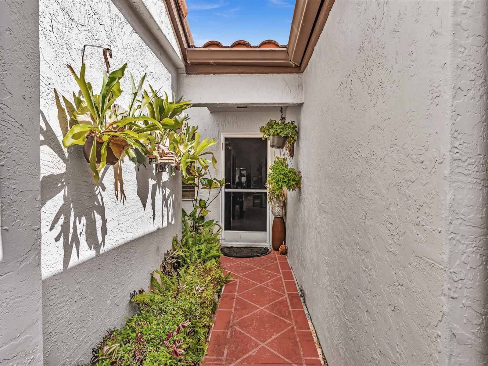 A narrow outdoor entryway features a red tiled path, white stucco walls, and hanging plants — 6454 Pointe Pleasant Circle, Delray Beach FL 33484, DELRAY BEACH, FL