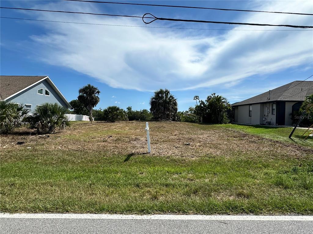 A vacant, grassy residential lot sits between two existing houses under a blue sky — 17063 OHARA DRIVE, PORT CHARLOTTE, FL