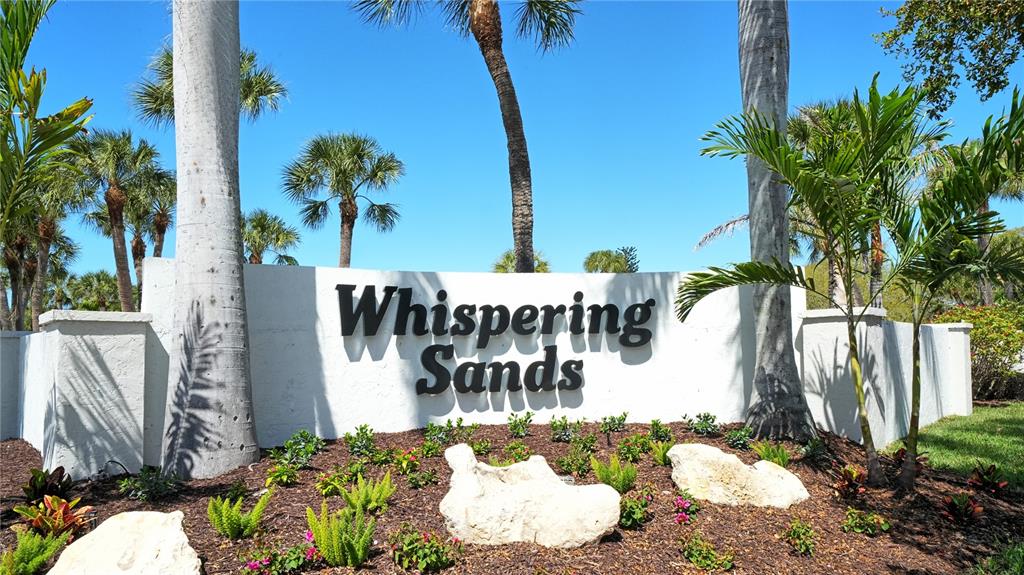 A white stucco sign reading Whispering Sands sits in a garden with palm trees — 199 WHISPERING SANDS DRIVE UNIT 204, SARASOTA, FL