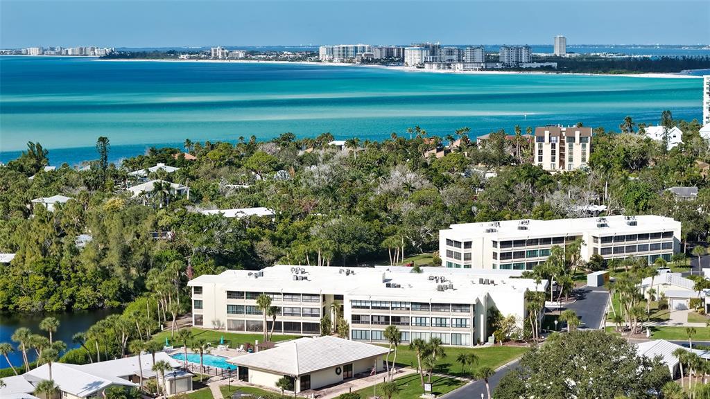 An aerial view shows low-rise apartment buildings surrounded by lush trees near a turquoise bay — 199 WHISPERING SANDS DRIVE UNIT 204, SARASOTA, FL
