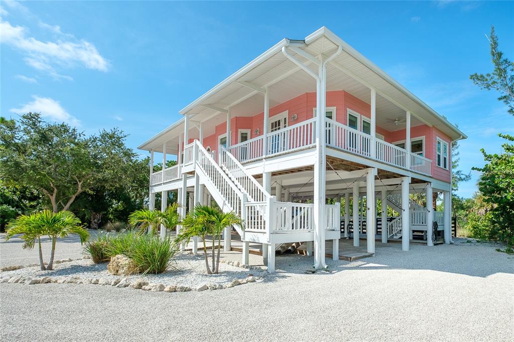 A pink stilt house with a white wraparound porch and stairs on a gravel lot — 141 GASPARILLA WAY, PLACIDA, FL