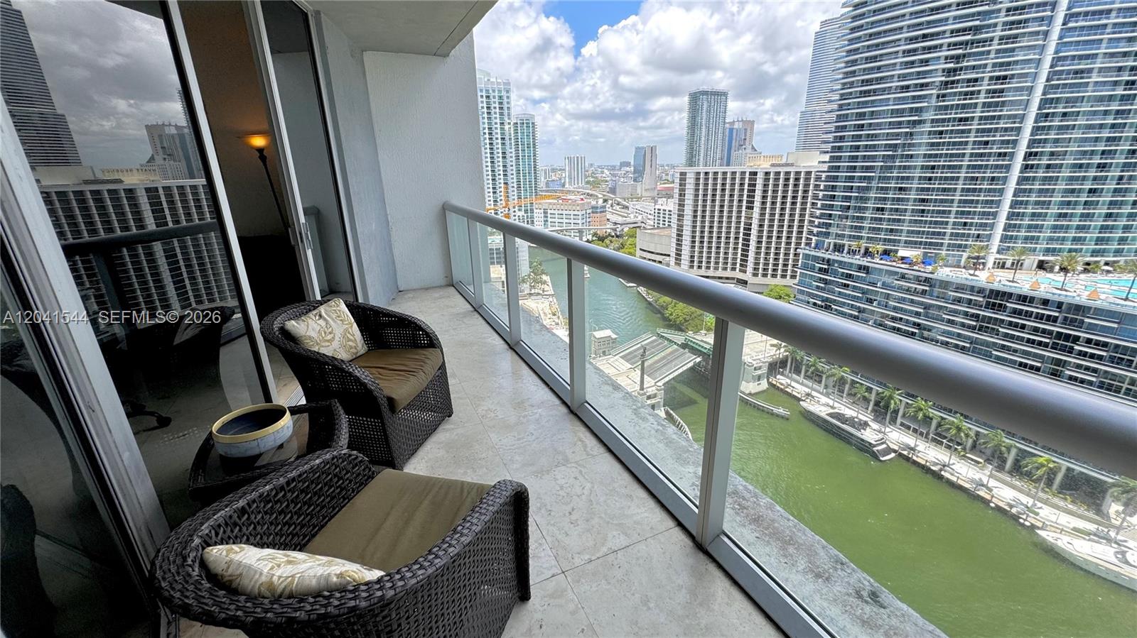 A balcony with wicker chairs overlooks a river, a drawbridge, and surrounding city skyscrapers — 475 Brickell Ave # 2512, Miami FL 33131, MIAMI, FL