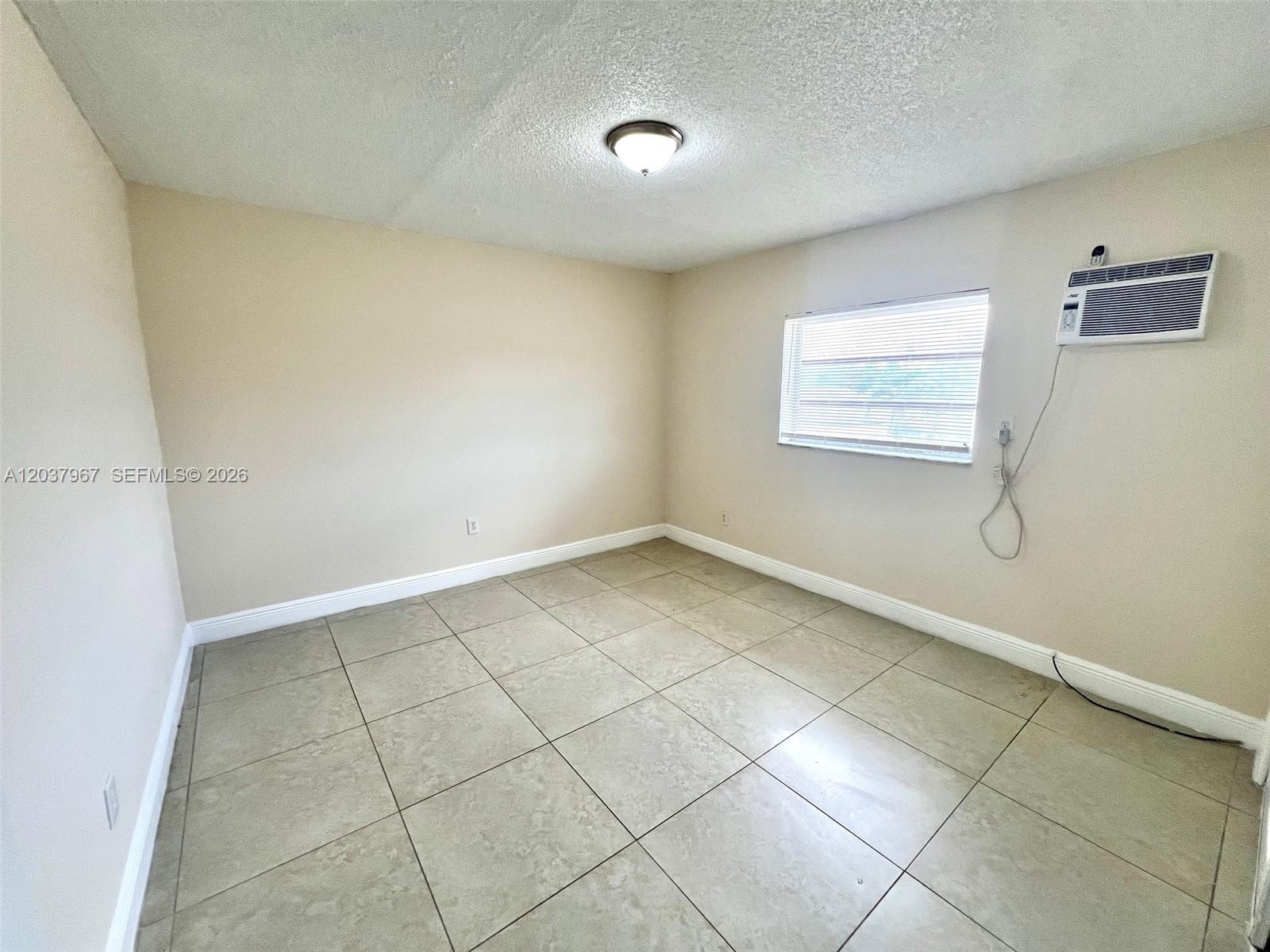 An empty room features beige walls, large light-colored floor tiles, a window, and an air conditioner — 685 NE 85th St # 9, Miami FL 33138, MIAMI, FL