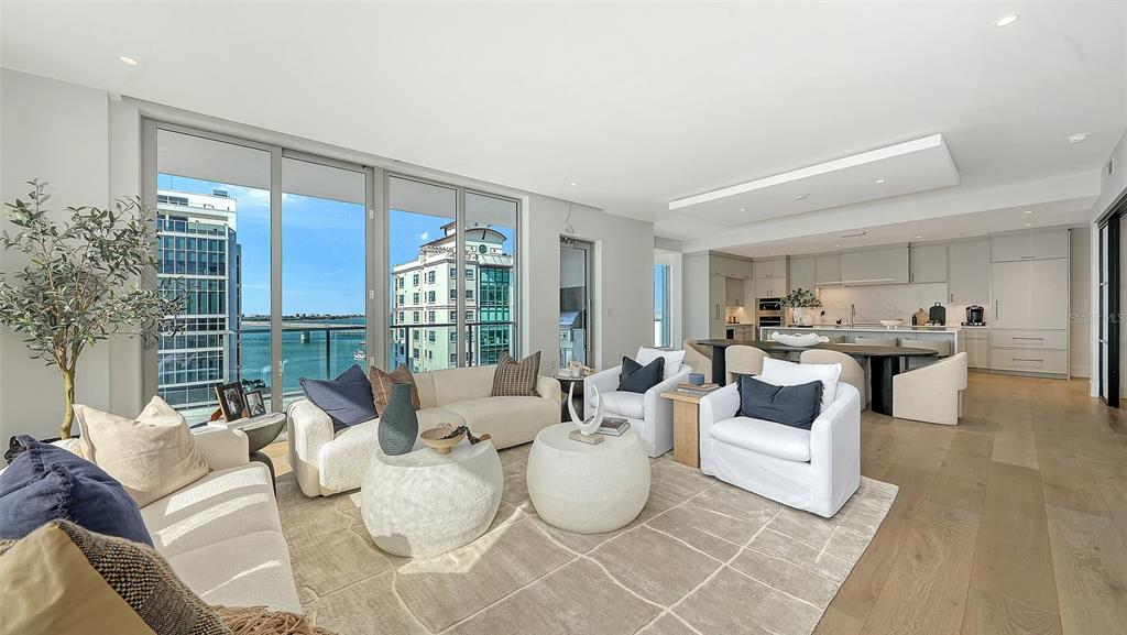 A modern living room with light wood floors, white furniture, and floor-to-ceiling windows overlooking water — 253 GOLDEN GATE POINT UNIT 703, SARASOTA, FL
