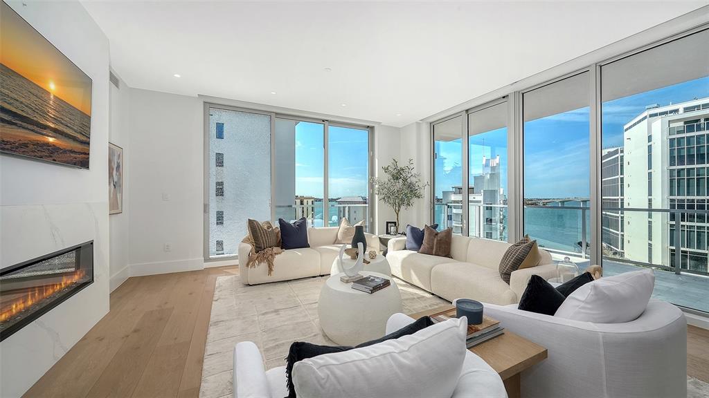 A modern living room features a fireplace, white sofas, and floor-to-ceiling windows overlooking water — 253 GOLDEN GATE POINT UNIT 703, SARASOTA, FL