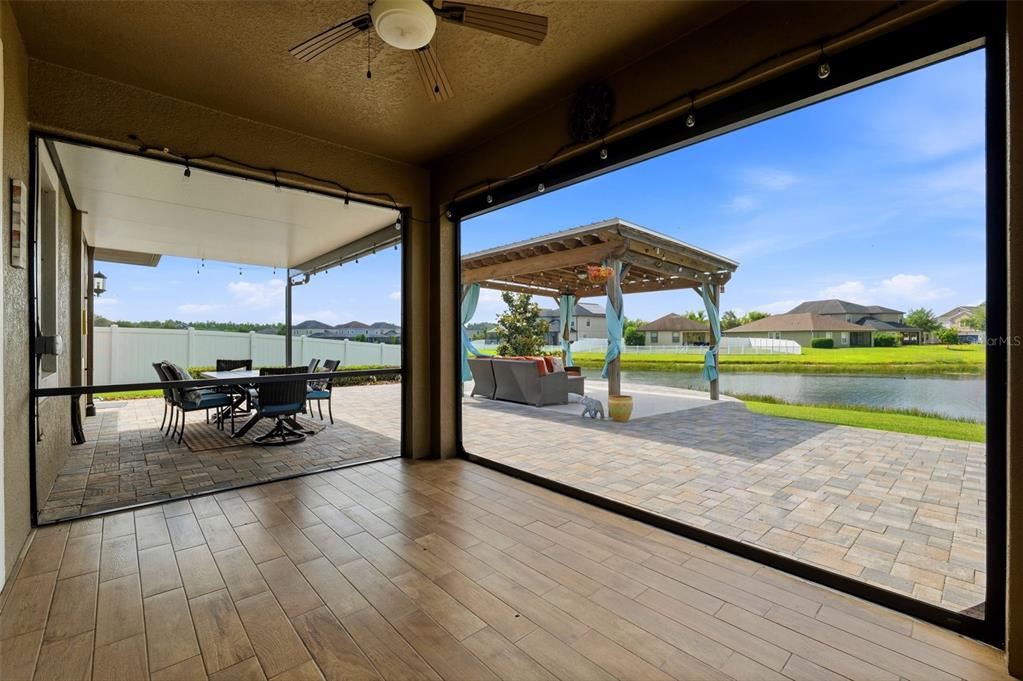 A covered patio with wood-look tile flooring overlooks a paved backyard, pergola, and pond — 19419 PADDOCK VIEW DRIVE, TAMPA, FL