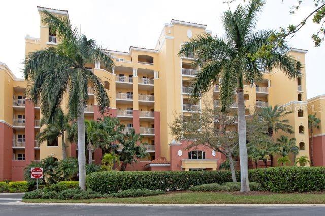 A multi-story yellow and red apartment building stands behind palm trees and a green hedge — 615 RIVIERA DUNES WAY UNIT 402, PALMETTO, FL