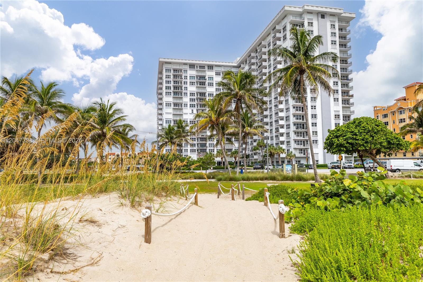 A sandy path leads toward a tall white apartment building surrounded by palm trees — 405 N Ocean Blvd # 117, Pompano Beach FL 33062, POMPANO BEACH, FL