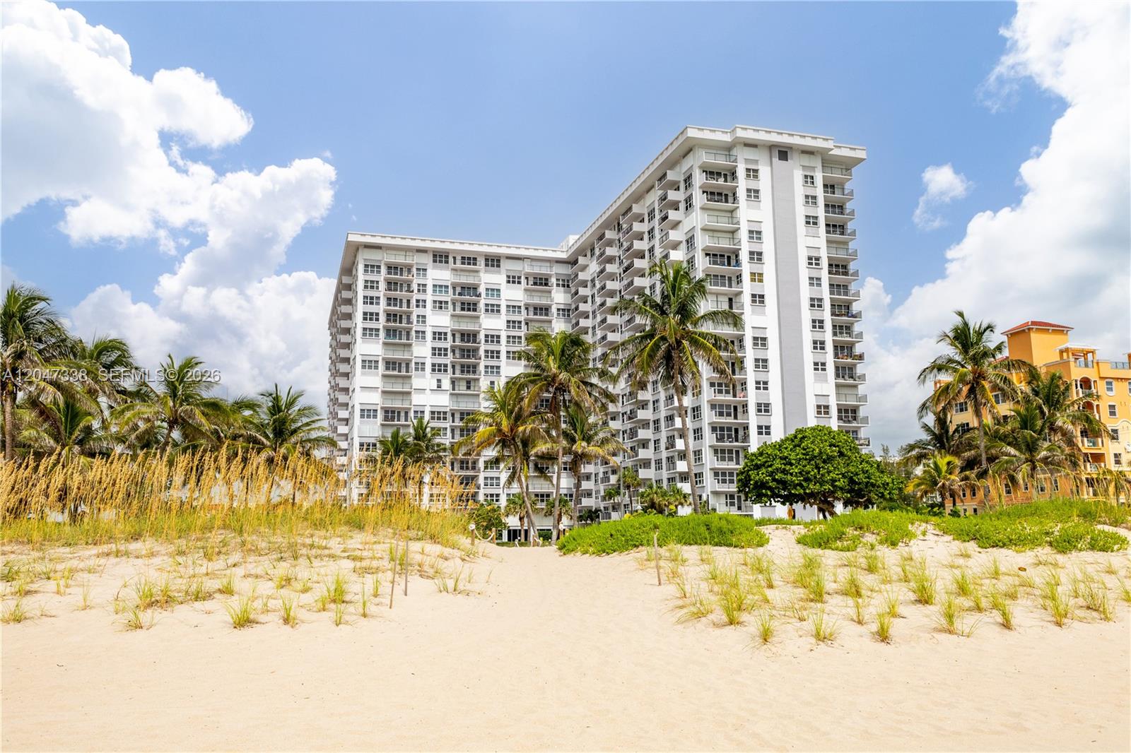 A tall white residential building stands behind a sandy beach with palm trees — 405 N Ocean Blvd # 117, Pompano Beach FL 33062, POMPANO BEACH, FL